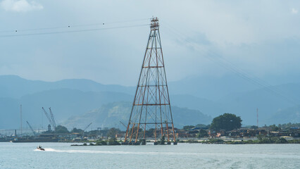 Large high voltage tower in a coastal city, with mountains in the background and a boat on the water