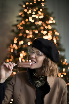 Cute young woman in winter outfit with berett, while biting into sweet holiday treats chocolate in front of Christmas tree. Funny fashionable woman enjoys eating sweets during X-mas