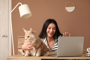 Pretty young woman with laptop and cute Maine Coon cat working near table at home