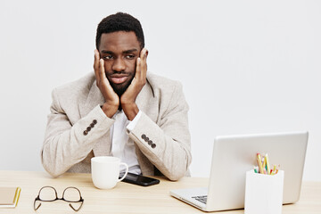 Frustrated young man with dark skin sits at desk with laptop, holding head in hands in discomfort....