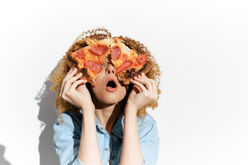 Young woman with curly hair expressing surprise while holding pizza slices, against a white...