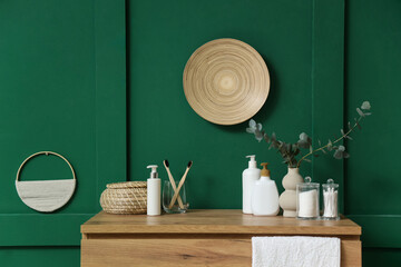 Vase with eucalyptus branches and cosmetic products on drawers near green wall in bathroom