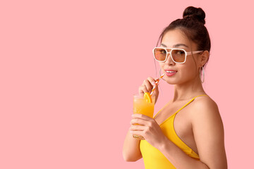 Pretty young woman in swimsuit with glass of orange juice and straw on pink background