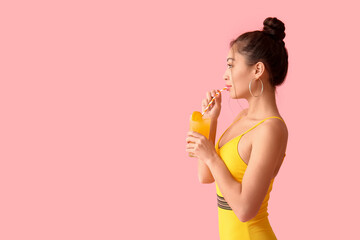 Pretty young woman in swimsuit drinking glass of orange juice with straw on pink background