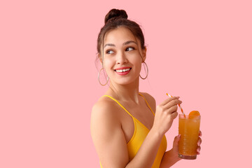 Pretty young woman in swimsuit holding glass of orange juice with straw on pink background