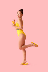 Happy young woman in swimsuit holding glass of orange juice with straw on pink background