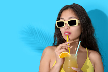 Pretty young woman in swimsuit and sunglasses holding glass of orange juice with straws on blue...