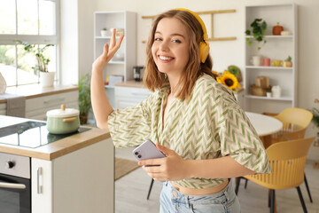 Cool young woman with mobile phone listening to music and dancing in kitchen