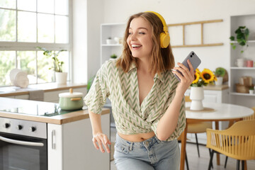 Cool young woman with mobile phone listening to music and dancing in kitchen