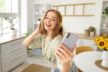 Cool young woman with mobile phone listening to music and dancing in kitchen