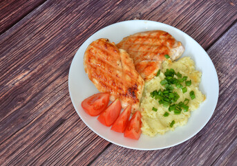 Grilled chicken fillet with mashed potatoes, green onions and tomatoes on a wooden table.