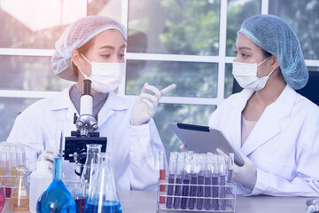 Two female scientists in lab coats and protective gear are performing an experiment, holding a flask with blue liquid while observing and taking notes on a tablet.