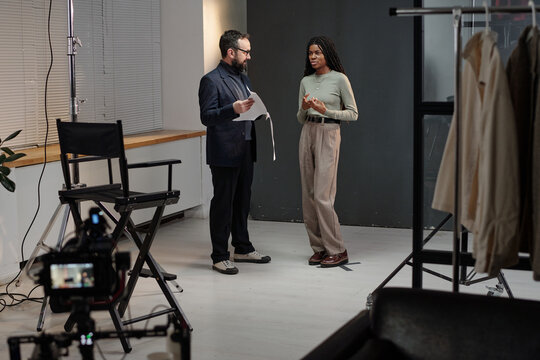 Caucasian man holding script talking with young adult Black woman during acting audition in studio setting, camera and director chair visible in foreground, clothing rack on side