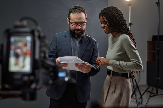 Caucasian man holding script and talking with young adult Black woman during acting audition in professional studio with camera and lighting equipment visible in foreground
