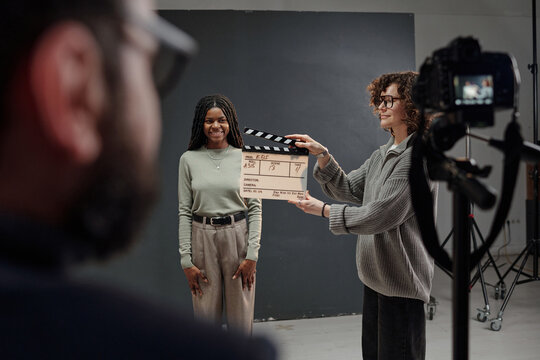 Black girl standing and smiling during acting audition while Caucasian woman holding clapperboard in front of camera, director observing scene