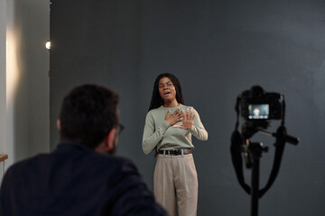 Black young adult woman performing monologue during acting audition, standing in front of camera with casting director observing, gesturing expressively with hands
