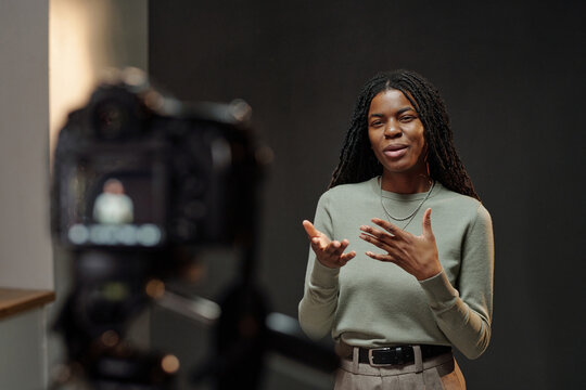 Young adult Black woman standing in front of camera gesturing with hands and speaking during acting audition, facial expression showing engagement, dark background visible
