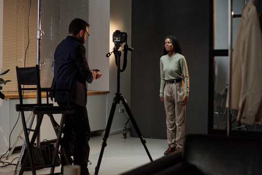 Black young adult woman standing in front of camera auditioning for acting role while Caucasian mature man directing scene in professional studio setting
