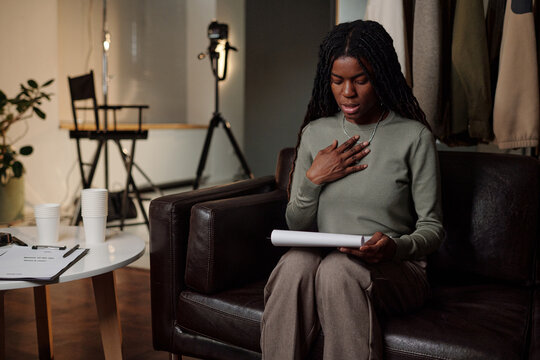 Black young adult woman sitting on sofa rehearsing script for acting audition, holding paper in one hand and touching chest with other hand, camera equipment visible in background