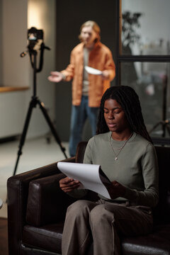 Black young adult woman sitting on sofa reading script during acting audition while Caucasian young adult man standing in background gesturing and holding script near camera setup