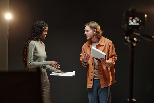 Young adult Black woman and young adult Caucasian man rehearsing script during acting audition, both holding papers, standing on stage with professional camera recording scene