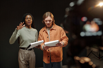 Young adult Black woman and young adult Caucasian man standing together holding scripts, rehearsing lines during acting audition in studio with professional camera visible in foreground