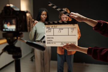 Young Black woman and young Caucasian man standing in front of camera during acting audition, holding script while crew member holding clapperboard in foreground, studio background visible