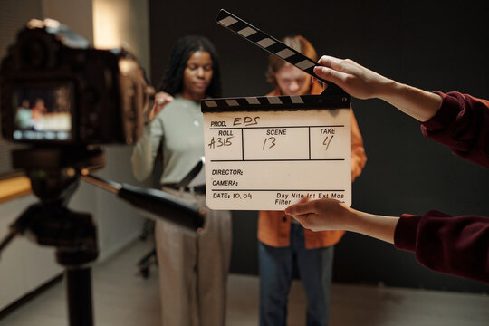 Young Black woman and young Caucasian man standing in front of camera during acting audition, holding script while crew member holding clapperboard in foreground, studio background visible