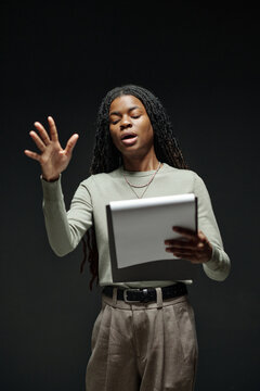 Young adult Black woman standing and performing dramatic monologue during acting audition, holding script in one hand and gesturing expressively with other hand against dark background