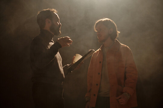 Caucasian middle aged man holding clipboard instructing Caucasian young adult man during acting audition on stage with dramatic lighting, both standing and interacting closely