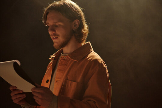 Young adult Caucasian man rehearsing script during acting audition, standing under stage lighting, holding papers and reading lines with focused expression, background in shadow
