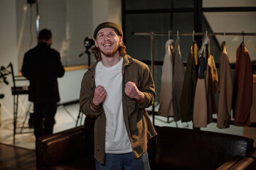 Caucasian young adult man standing in front of clothing rack clenching fists and smiling during acting audition while camera operator working in background studio environment
