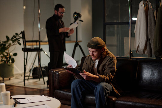 Caucasian young adult man sitting on sofa reading script during acting audition while middle aged Caucasian man standing in background holding papers near camera equipment