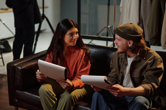 Young adult Caucasian man and young adult Hispanic woman sitting on sofa holding scripts, engaging in conversation during acting audition, professional studio equipment visible in background