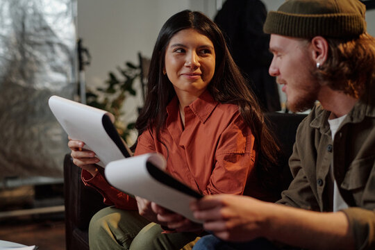Young adult Caucasian man and young adult Hispanic woman sitting together holding scripts, preparing for acting audition, engaging in conversation and exchanging expressions