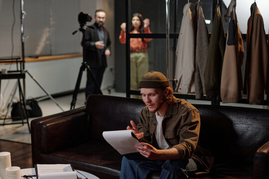 Young adult Caucasian man sitting on sofa reading script during acting audition, camera and tripod in background, two people standing behind glass observing scene