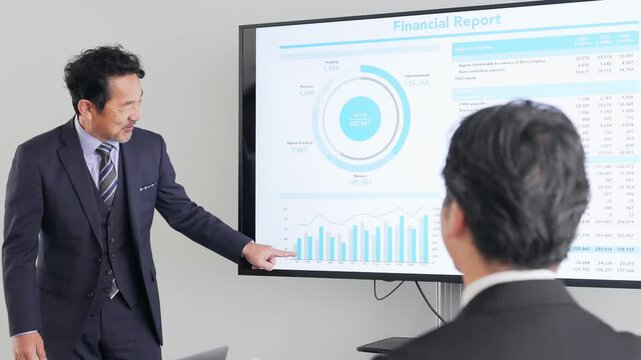 A middle-aged male business person giving an explanation in front of a large screen monitor