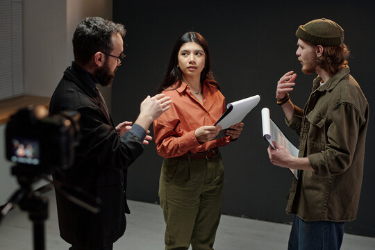 Young adult Caucasian man and young adult Hispanic woman holding scripts standing with middle aged Caucasian man gesturing during acting audition in studio setting