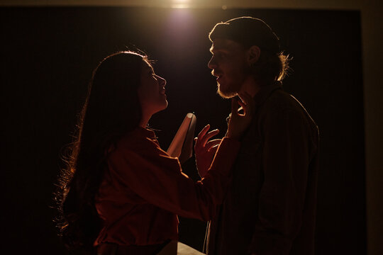 Young woman holding script standing close to man during acting audition, woman touching man's chin, both facing each other under dramatic lighting