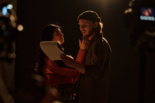 Young adult Caucasian man and Hispanic woman performing dramatic scene during acting audition, man holding script while woman touching his face, stage lights and cameras visible