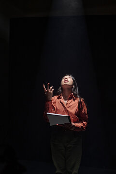 Hispanic woman standing under spotlight holding script looking upward performing dramatic monologue during acting audition on dark stage background