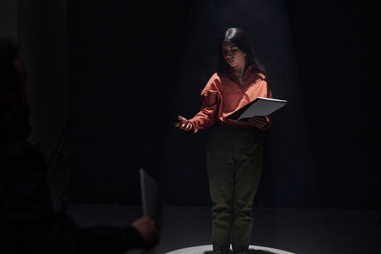 Young adult Hispanic woman standing on stage holding script, performing monologue during acting audition, gesturing with hand while looking at casting director in dark theater setting