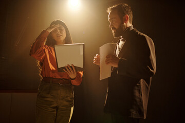 Young adult Hispanic woman holding script and shielding eyes from light while standing next to young adult Caucasian man holding papers during acting audition on stage