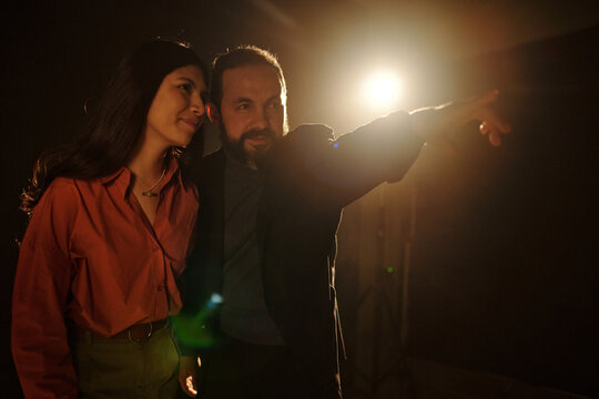 Hispanic woman standing beside Caucasian middle aged man pointing forward, both smiling and looking in same direction during acting audition on dark stage with bright spotlight
