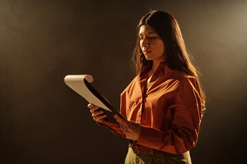Hispanic woman standing on stage holding script reading lines during acting audition focused on text with serious expression under dramatic lighting