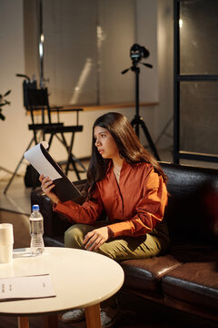 Hispanic woman sitting on sofa holding script and rehearsing lines during acting audition, focused expression on face, film equipment and director chair visible in background
