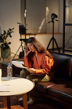 Hispanic woman sitting on sofa reading script and preparing for acting audition in studio setting with professional camera equipment visible in background