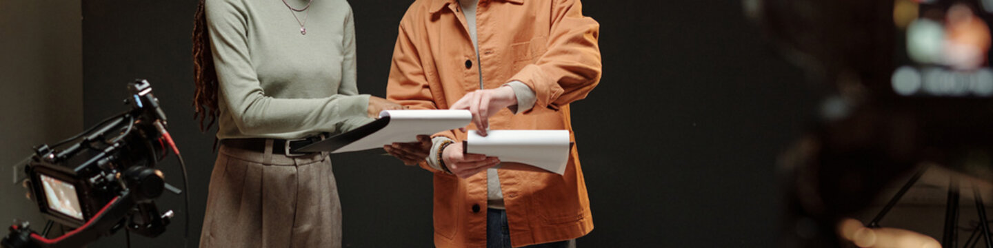 Young adult Black woman and young adult Caucasian man standing together holding scripts, rehearsing lines during acting audition with professional video camera equipment visible in foreground