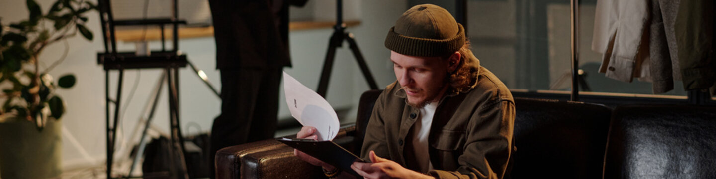 Caucasian young adult man sitting on sofa reading script and preparing for acting audition, holding papers and focusing on lines while another stands in background near equipment