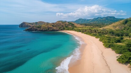 Panoramic aerial view of a pristine beach with turquoise water, light beige sand, lush green vegetation, and rolling hills in the background under a partly cloudy sky.